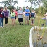 Group of students attentively listening to an instructor by a small outdoor pond.