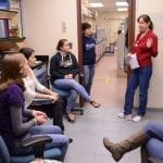 A group of focused students listening to a woman speaking and gesturing, possibly during a laboratory or educational briefing.