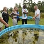 Group of students learning about aquatic ecosystems from an instructor beside an outdoor pool.