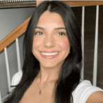 Person with long dark hair smiling in a selfie, standing indoors near a wooden staircase, wearing a white top and pendant necklace.