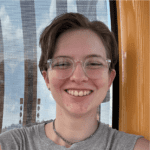 A person with short hair and glasses smiles at the camera, wearing a gray sleeveless top and a chain necklace, with a patterned background.