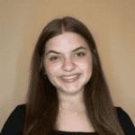 A smiling person with long brown hair, wearing a black top, stands against a plain beige background.