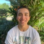 Person smiling outdoors, wearing a white T-shirt with graphics, surrounded by green plants.