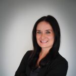 A woman with long dark hair, wearing a black blazer and gray top, stands with arms crossed and smiles at the camera against a plain background.