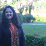 A woman with long dark hair, wearing a black jacket and a colorful scarf, stands outdoors with greenery and trees in the background.