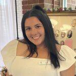 A woman with long dark hair wearing a white top stands indoors, smiling at the camera. Behind her is a bulletin board with photos and flowers.