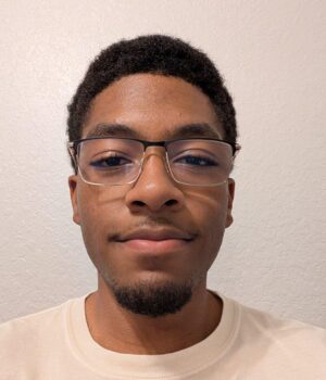 A young man with short curly hair, glasses, and a trimmed beard is wearing a light-colored shirt and standing against a plain, textured light wall.