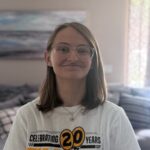 Person with shoulder-length brown hair and glasses, wearing a "Celebrating 20 Years" t-shirt, standing indoors with a blurred background.