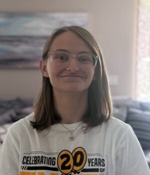 Person with shoulder-length brown hair and glasses, wearing a "Celebrating 20 Years" t-shirt, standing indoors with a blurred background.