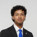 A person with short curly hair wearing a black suit, white shirt, and blue tie, sitting against a plain light background.