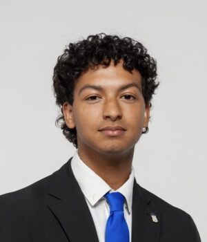 A person with short curly hair wearing a black suit, white shirt, and blue tie, sitting against a plain light background.