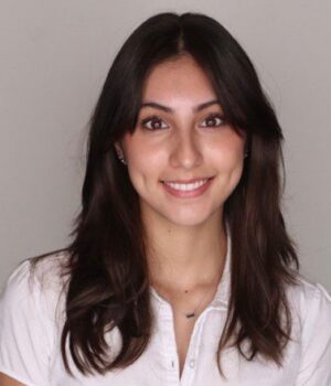 A young woman with long dark hair, wearing a white short-sleeve blouse, smiles at the camera against a plain light gray background.
