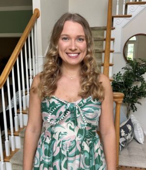A woman with long curly hair in a green patterned dress stands smiling in front of a staircase inside a well-lit home.