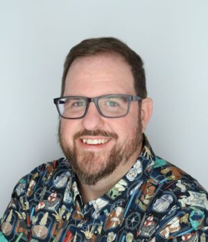 A man with glasses and a beard smiles at the camera, wearing a colorful shirt with cartoon character designs and a smartwatch.